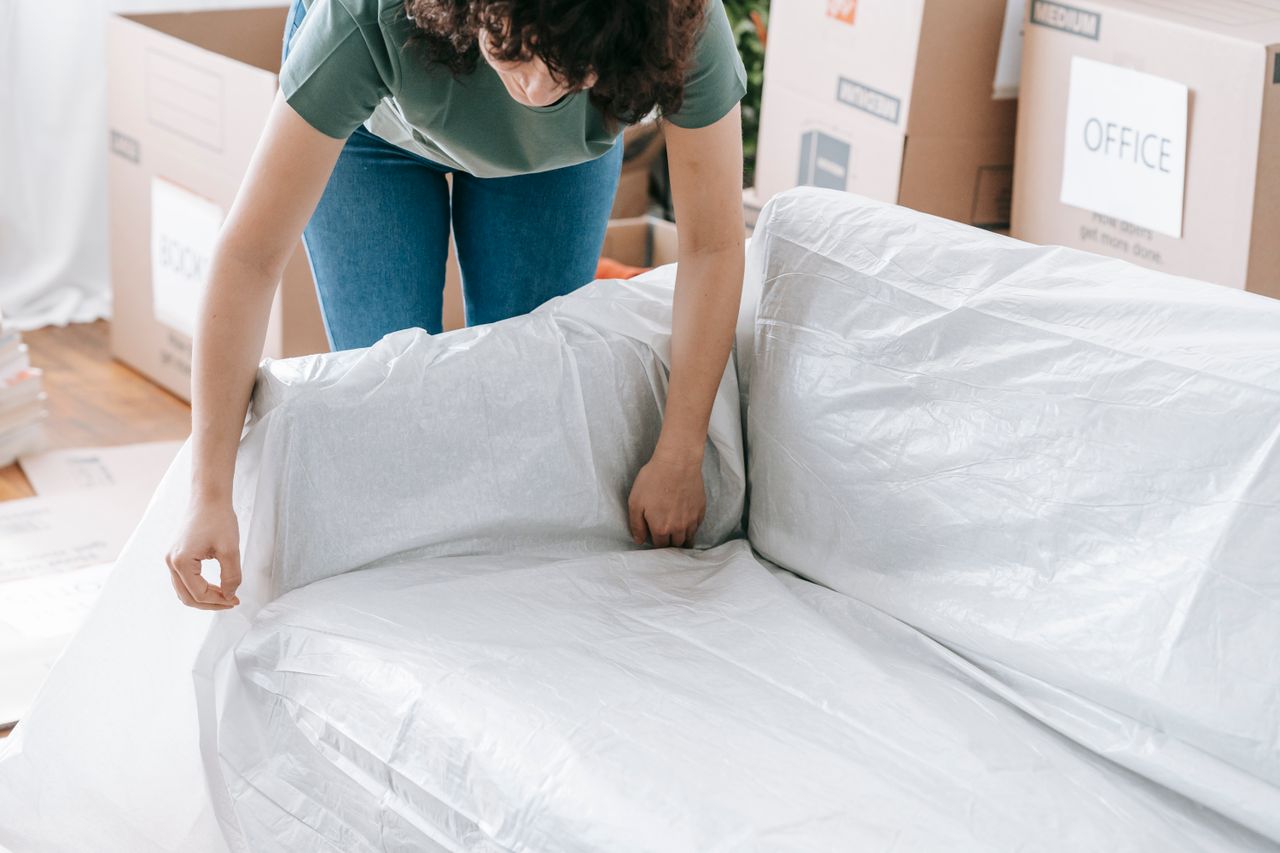 Une femme enveloppe soigneusement un canapé dans un matériau de protection tout en emballant des cartons dans une pièce lumineuse.